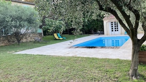 Una piscina con solárium de piedra, un edificio pequeño y un tobogán en un patio trasero.
