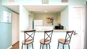 Une cuisine avec un mur bleu clair, un réfrigérateur blanc, un four à micro-ondes et une machine à café noire. On y trouve trois tabourets de bar au design à motifs croisés.