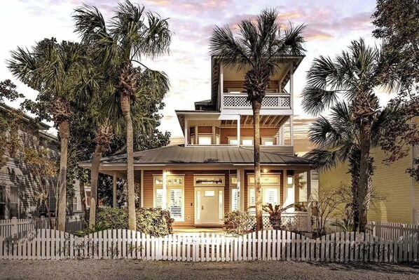 A two-story house with a balcony, surrounded by a white picket fence and palm trees.