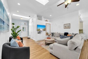 A modern living room with a grey sofa, a black armchair, and a coffee table. There is a television mounted on the wall and a ceiling fan above. - Timeless Traditions-Golf Cart Deals-Steps to Beach (Destin)