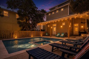 A swimming pool with lounge chairs and string lights, surrounded by a wooden deck and a building with multiple windows.