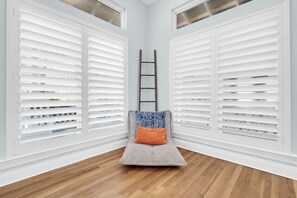 A room with a wooden floor, white shutters, a ladder, and a cushioned bench with patterned cushions.