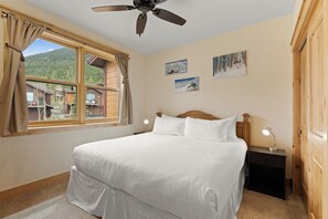 Une chambre à coucher avec un lit, un ventilateur de plafond, une table de chevet avec une lampe, une fenêtre avec des rideaux et des tableaux accrochés au mur.