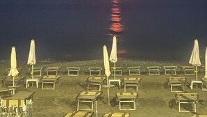Una playa con filas de tumbonas y sombrillas, el sol poniéndose sobre el agua y un mar tranquilo.
