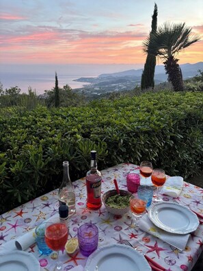 An outdoor dining table set with drinks and a bottle, overlooking a scenic view of the sea and mountains.