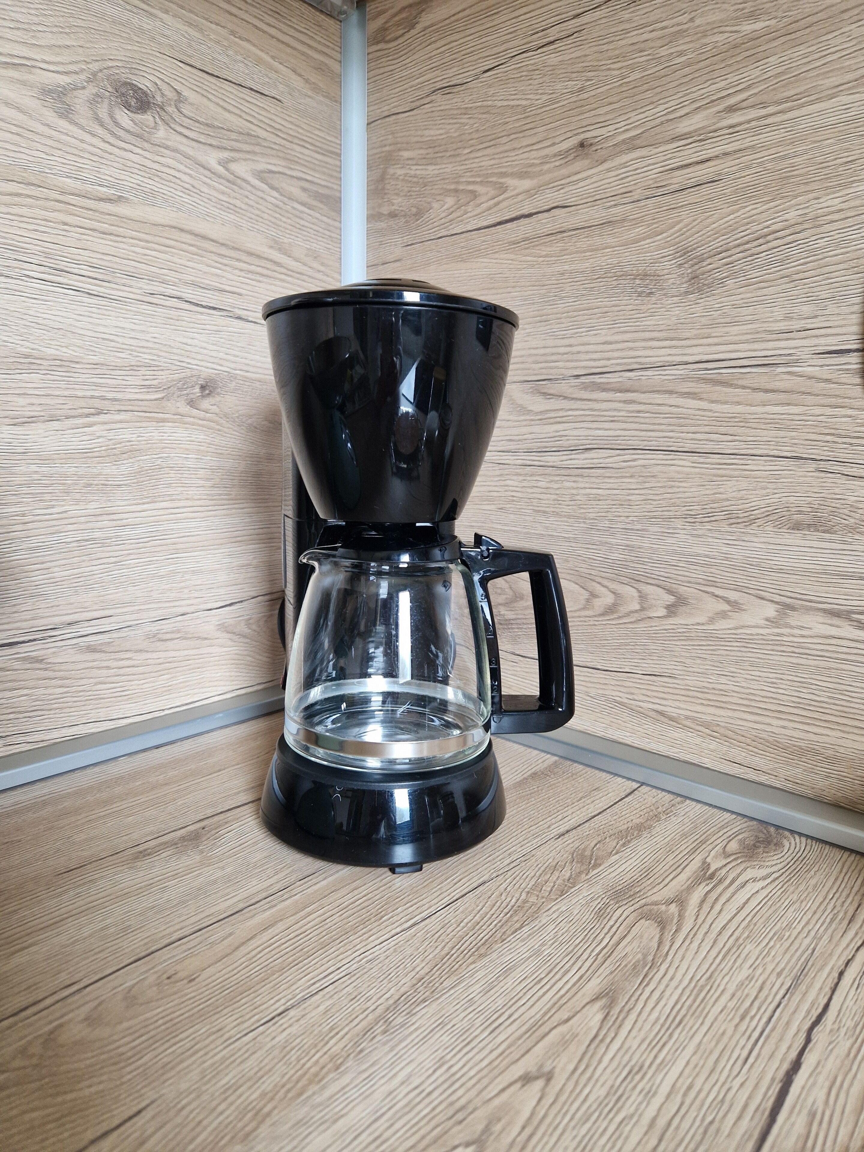 A black coffee maker with a clear carafe and a handle, placed on a wooden floor.