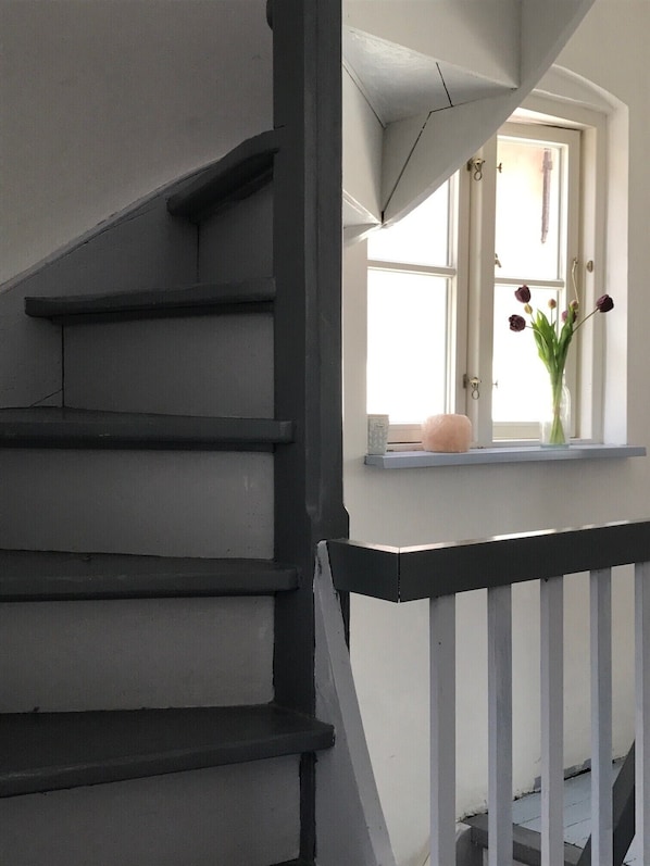 Una escalera con barandillas negras y paredes blancas que conduce a una plataforma con una ventana y un jarrón de flores.