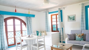 A bright living room with a white sofa, a round table, and a white chair. There is a window with blue curtains and a door with a wooden frame.