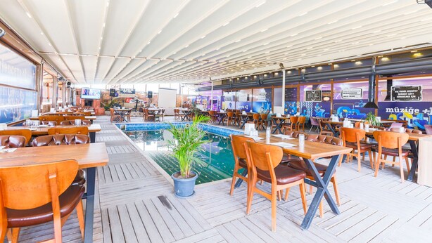 A spacious outdoor dining area with wooden tables and chairs, a central pool, and a covered ceiling. - İznik Otel (Iznik)