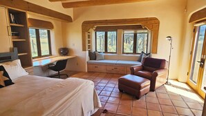 A bedroom with a large bed, a desk, a chair, and a window seat with cushions. - Sweeping panoramic mountain views, expansive adobe privacy wall system (Arroyo Seco)