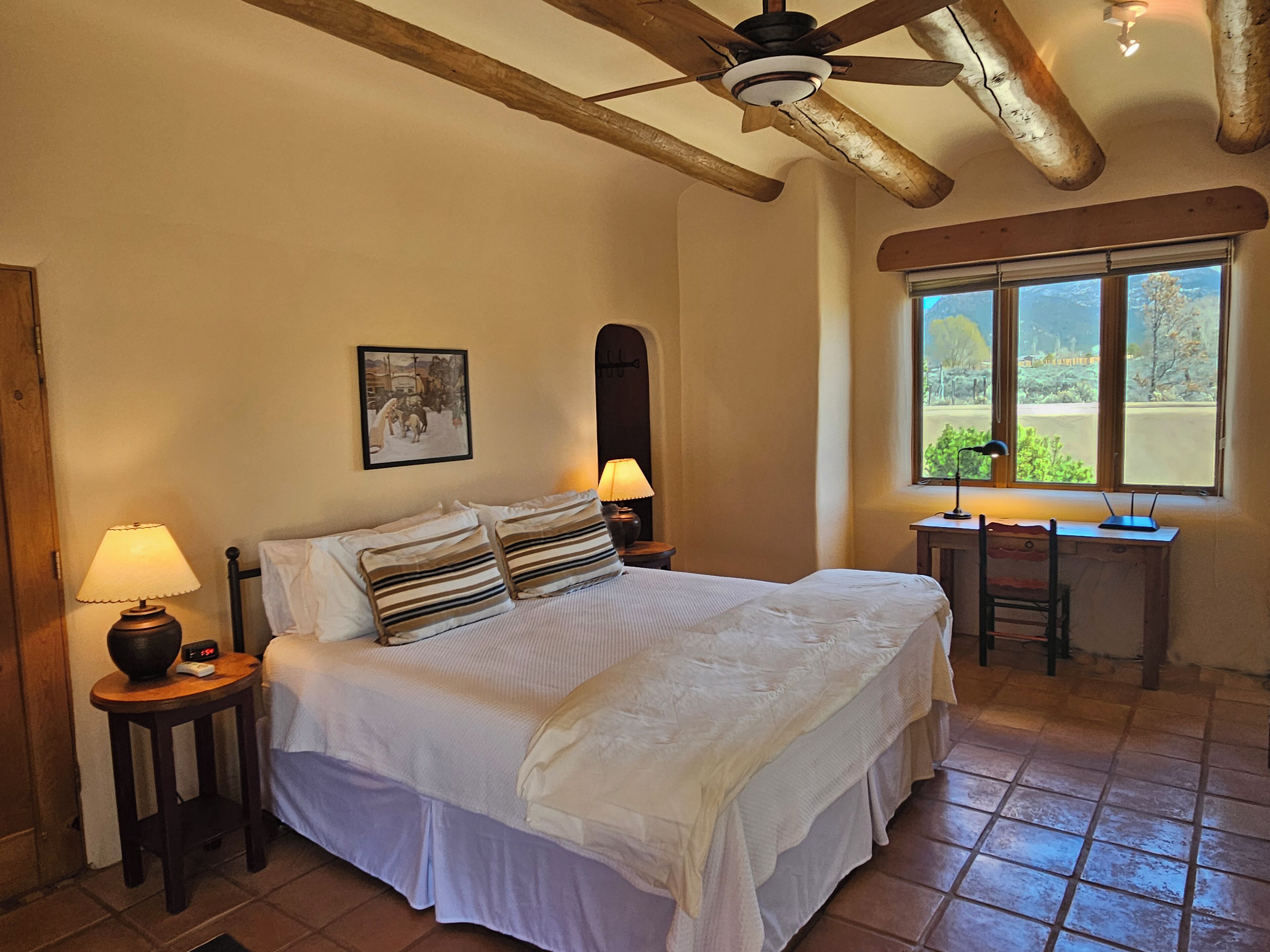 A bedroom with a bed, two bedside lamps, a desk, and a window with a view of greenery.