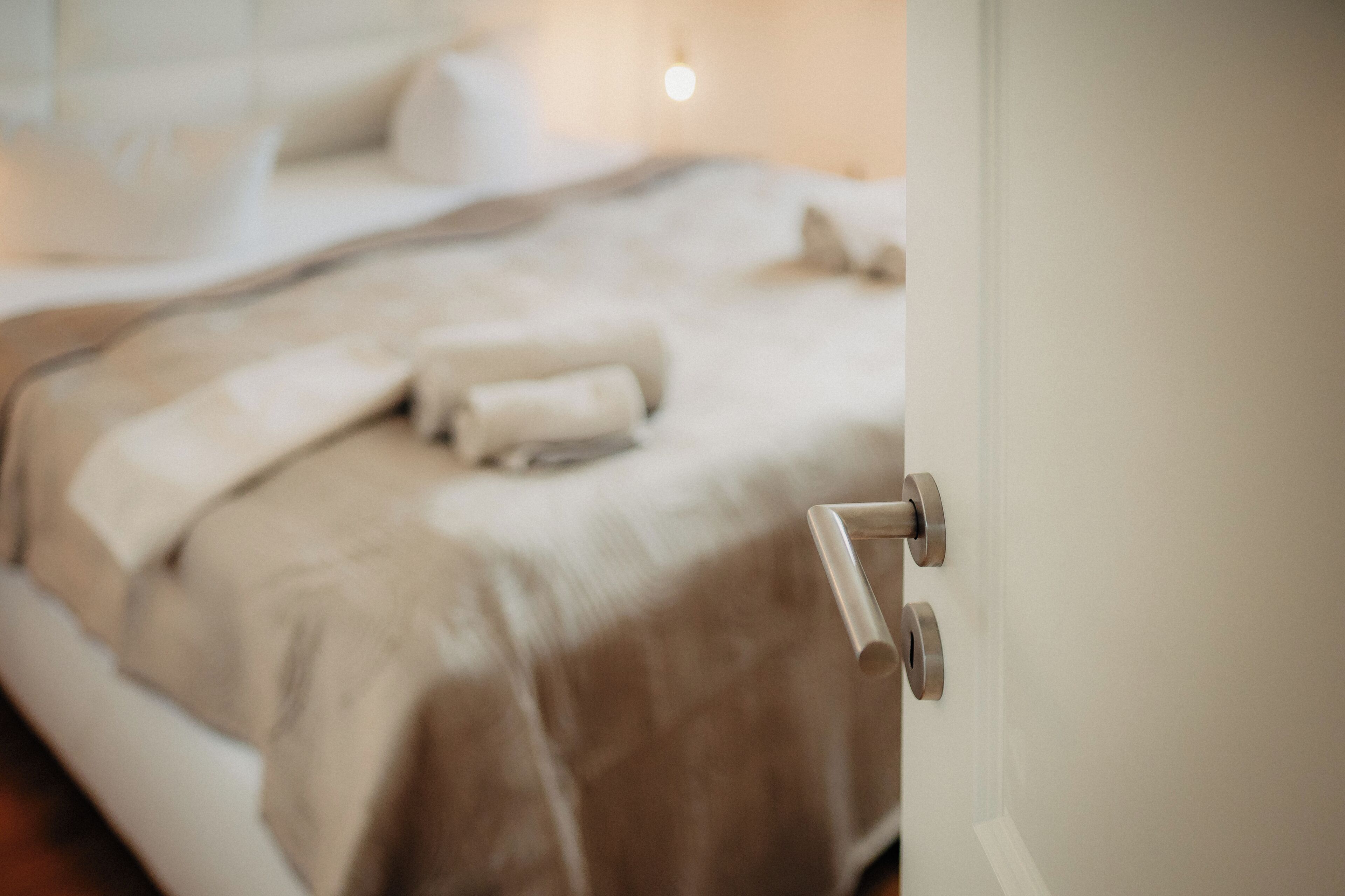 Una cama con una colcha color beige y almohadas, una mesita de noche y una puerta con pomo plateado.