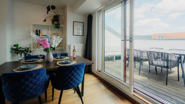 Un comedor con mesa de madera, sillas tapizadas en azul, un jarrón con flores y vista a una terraza a través de un ventanal.