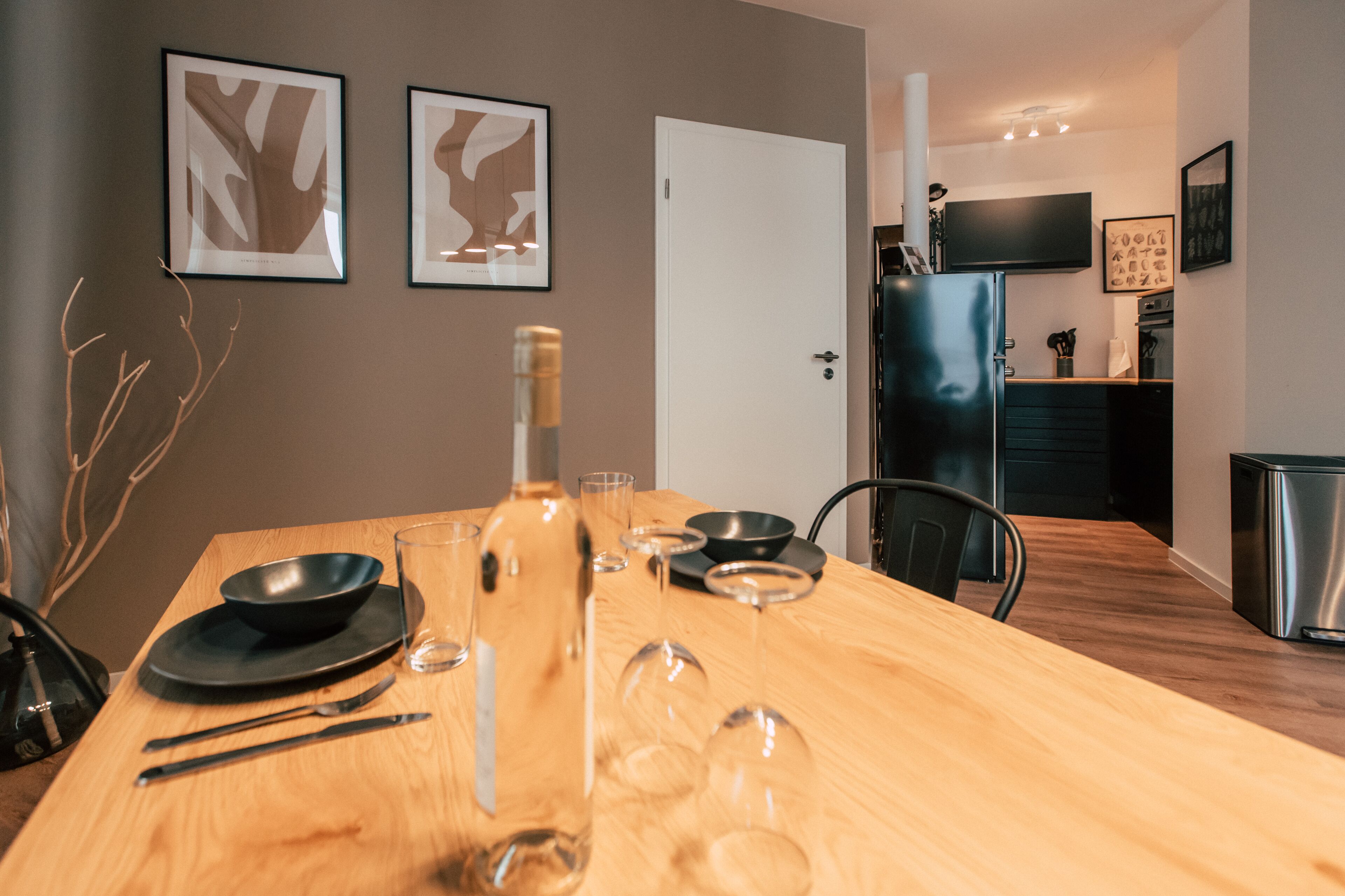 A dining table set with plates, cutlery, wine glasses, and a bottle, with a modern kitchen and a door in the background.