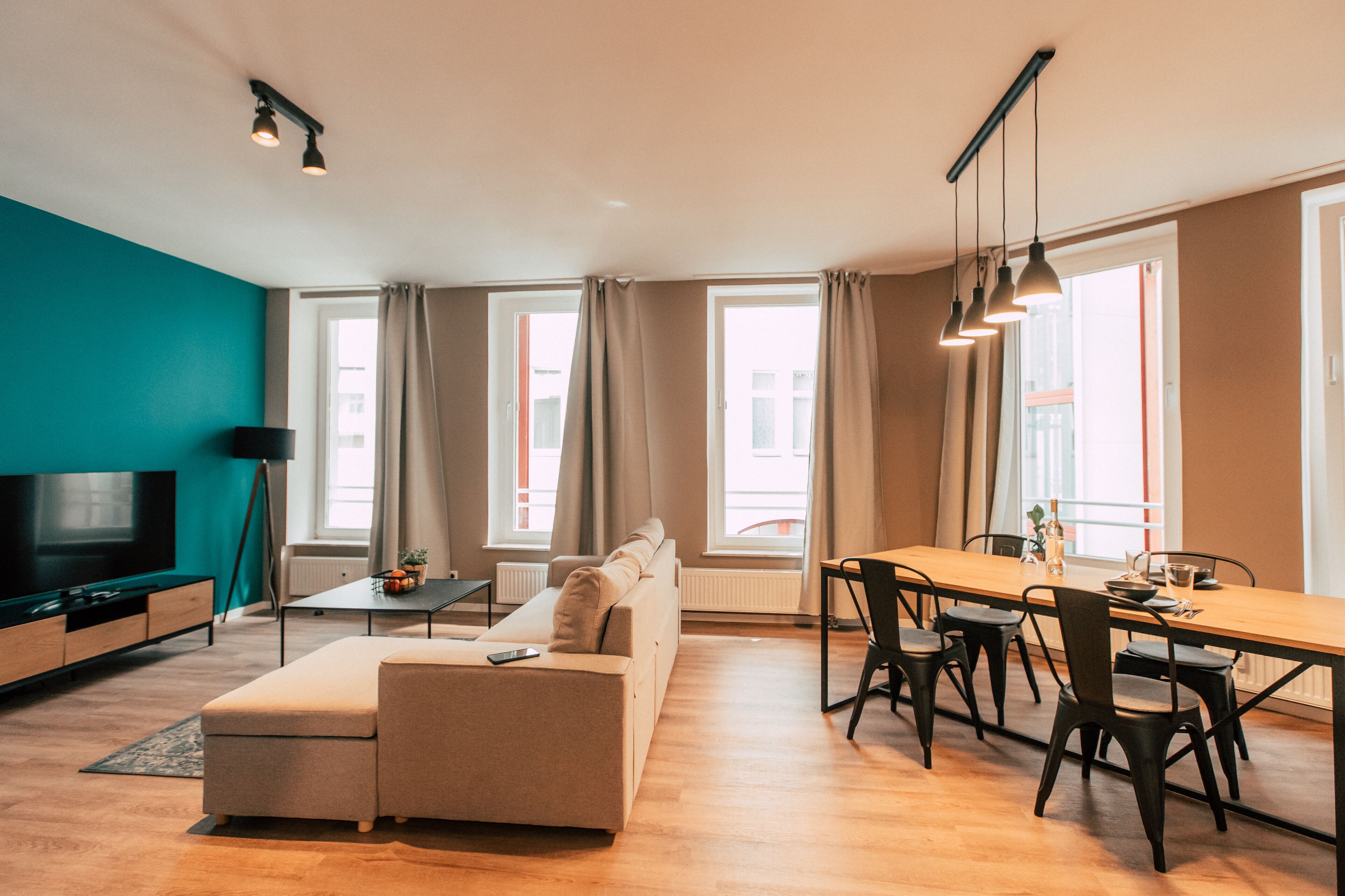 A modern living room with a sofa, a dining table with chairs, a TV, and large windows with curtains.