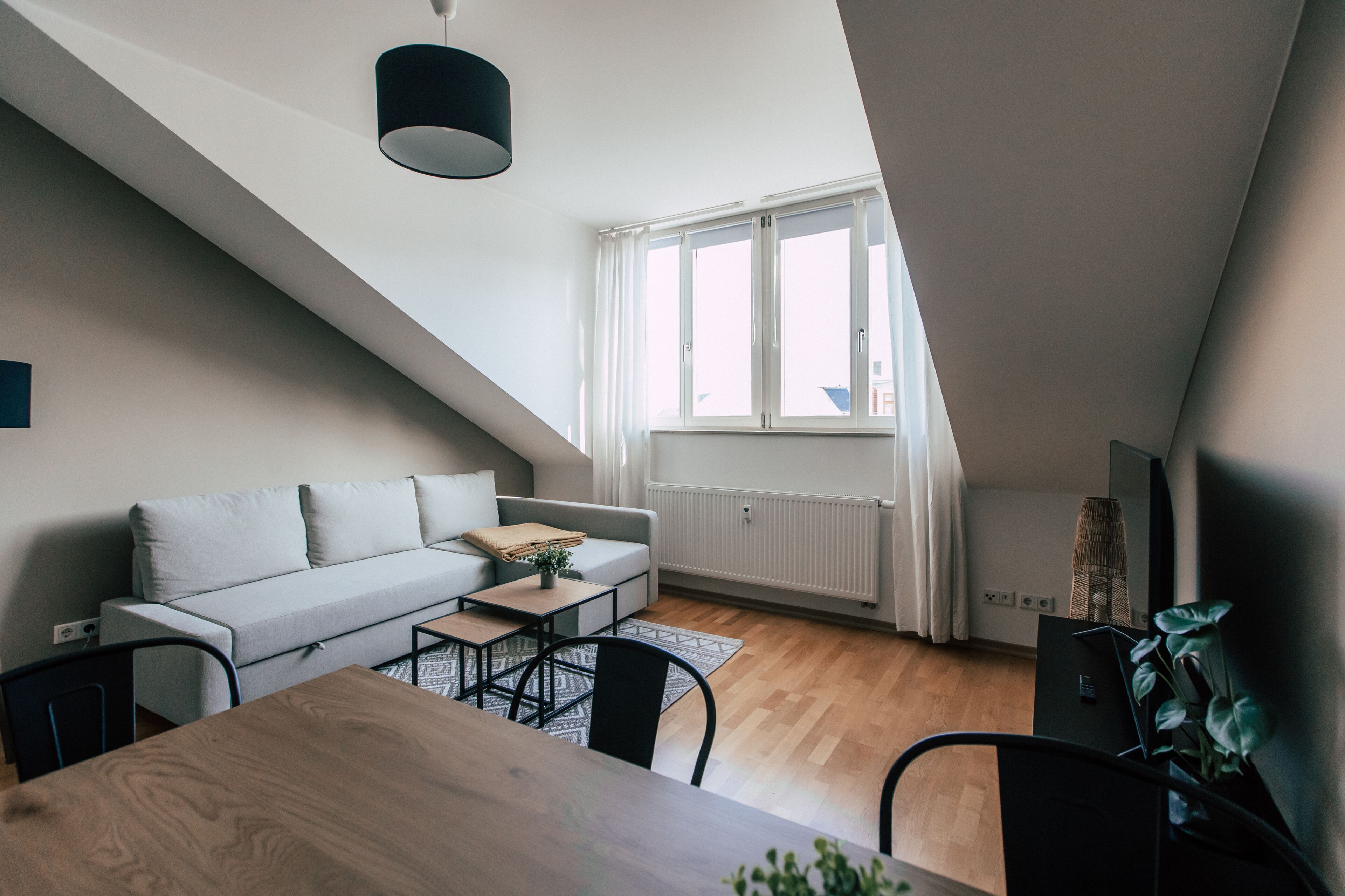 Ein modernes Loft-Apartment mit schrägverdachter Decke, einem Essbereich mit Holztisch und -stühlen, einem grauen Sofa und einem großen Fenster mit Vorhängen.