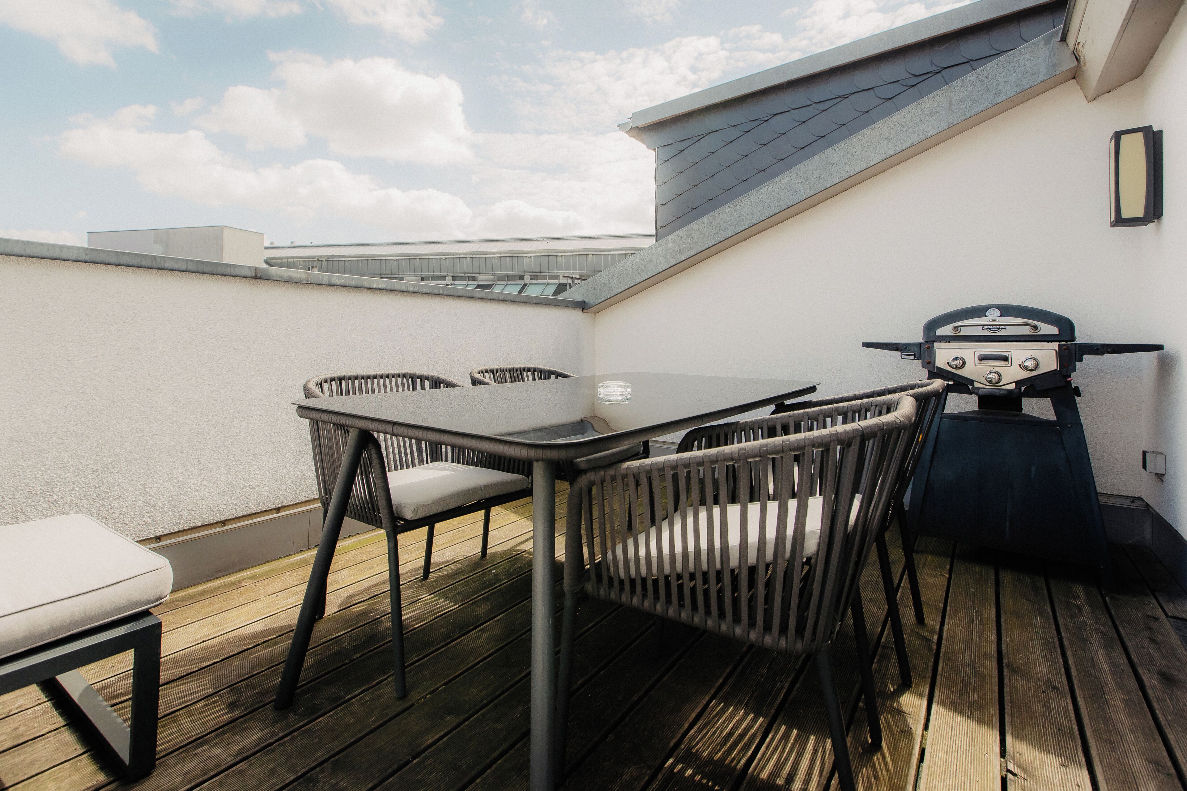 A rooftop terrace with a table and chairs, a grill, and a view of the sky.
