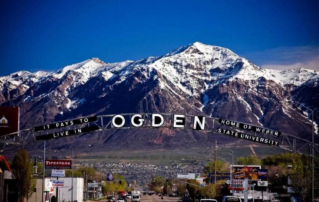 A mountain range with a sign reading "OGDEN HOME OF WEBER STATE UNIVERSITY" and a road leading into a town.