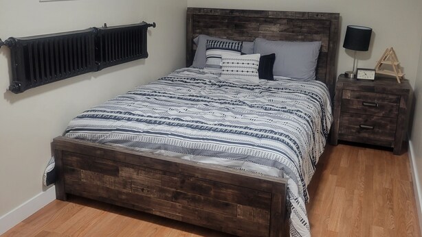 A bedroom with a wooden bed, a nightstand, a radiator, and two windows with blinds. - Beautiful Newly Renovated Basement Apartment (Ogden)