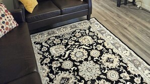A living room with a dark brown leather sofa, a patterned rug, a wooden dining table, and a kitchen area in the background. - Beautiful Newly Renovated Basement Apartment (Ogden)