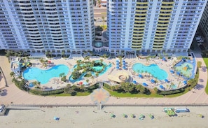 A high-rise hotel with multiple balconies, a central swimming pool area with multiple pools, and a landscaped garden.