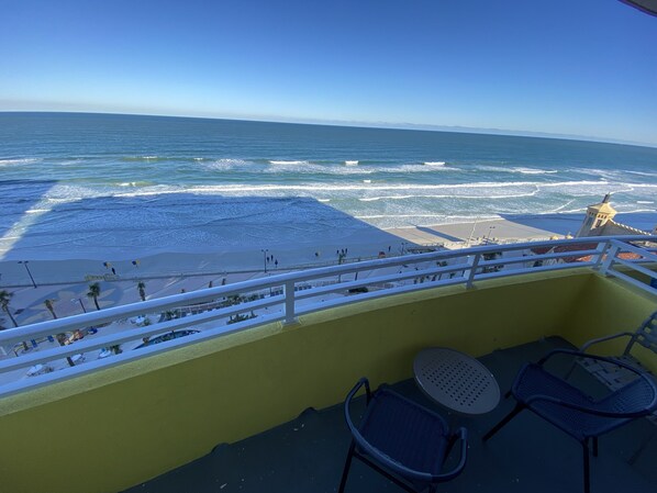 A balcony with a view of the beach and ocean. There are two chairs and a table on the balcony.
