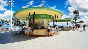 Un bar frente a la playa con una decoración en tonos verde y amarillo, situado en una zona pavimentada con palmeras y sillas de playa.