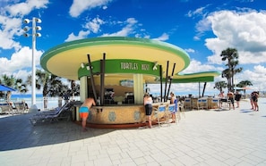 A beachside bar with a green and yellow color scheme, located on a paved area with palm trees and beach chairs.