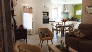 A living room with a sofa, armchair, and coffee table, a dining area with a table and chairs, and a kitchen with appliances and a green wall. - Gite Pouquette (Aramits)