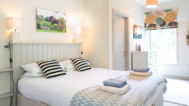 Ein ordentlich bezogenes Bett mit einem gestreiften Kopfkissen, einem gefalteten Handtuch und einer gemusterten Tagesdecke. Ein weißer Nachttisch mit einer Lampe, ein gerahmtes Bild an der Wand und ein Fenster mit einem Blumenrollo sind ebenfalls zu sehen.