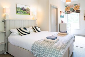 A neatly made bed with a striped pillow, a folded towel, and a patterned throw blanket. There is a white bedside table with a lamp, a framed picture on the wall, and a window with a floral blind.