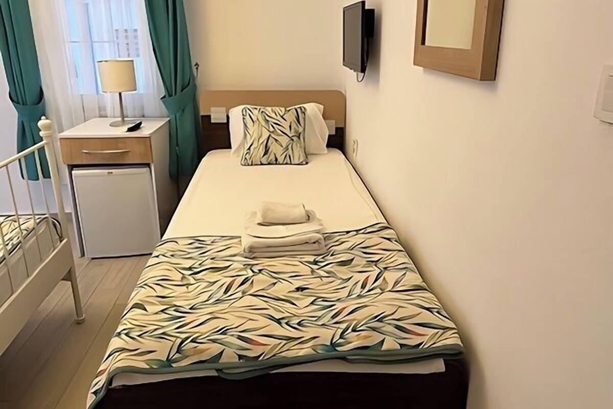 Une chambre d’hôtel avec un lit simple, une table de chevet avec une lampe, un téléviseur fixé au mur et une fenêtre avec des rideaux.