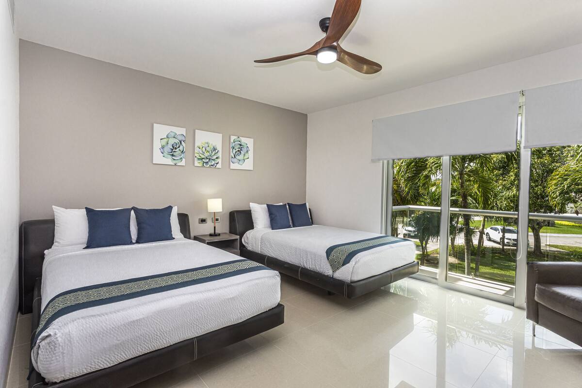 A modern hotel room with two beds, a ceiling fan, artwork on the wall, and a view of the outdoors.