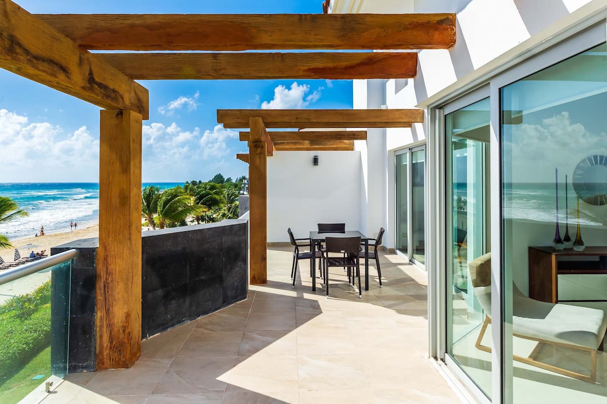 A balcony with a wooden pergola, a table with chairs, and a view of the beach and ocean.