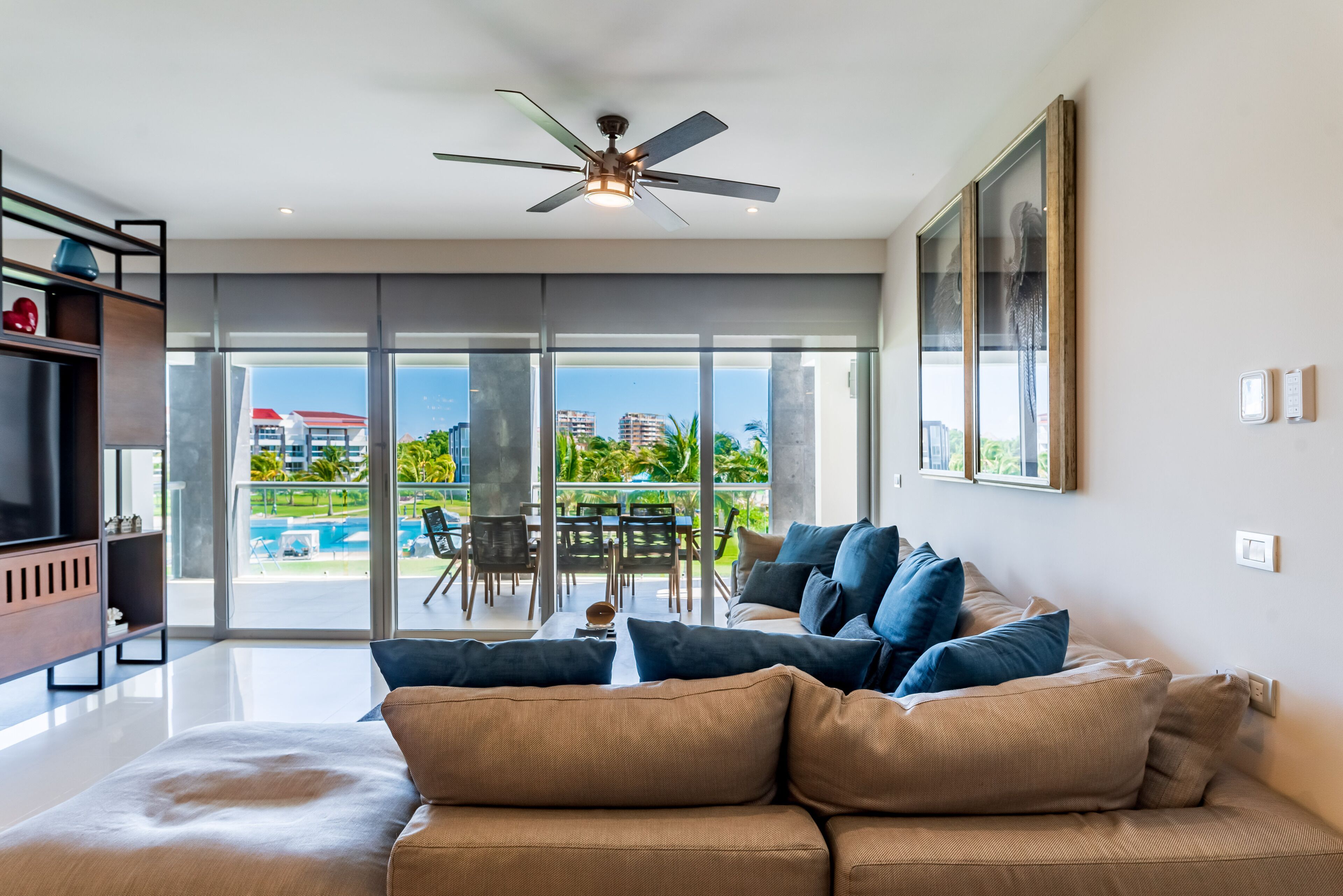 Un salon moderne avec un grand canapé, un ventilateur de plafond et une vue sur une piscine et des bâtiments à l’extérieur.