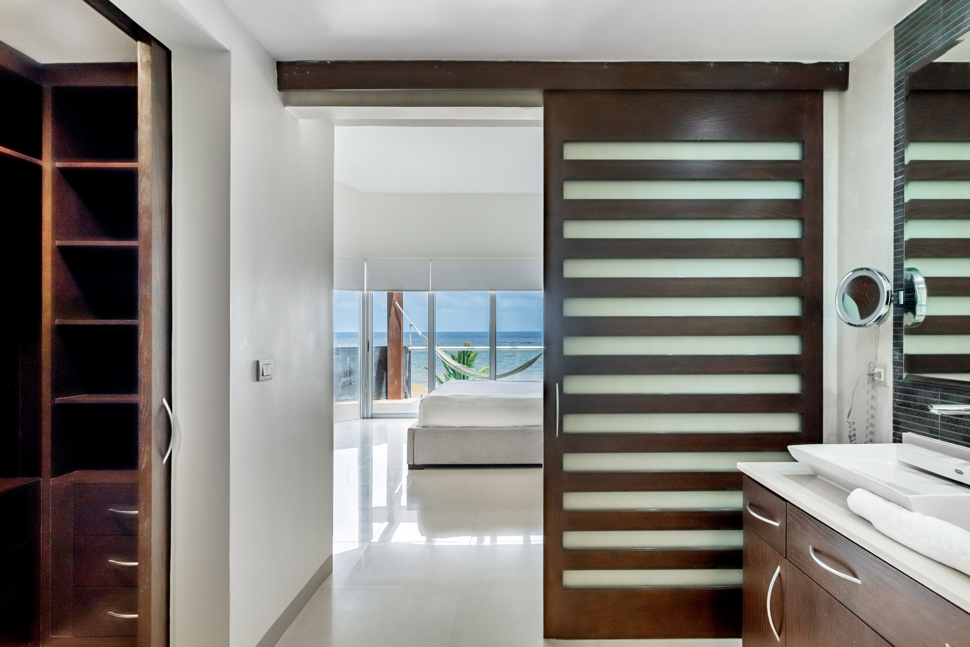 A modern bathroom with a large mirror, a wooden door with frosted glass panels, and a view of the ocean through large windows.