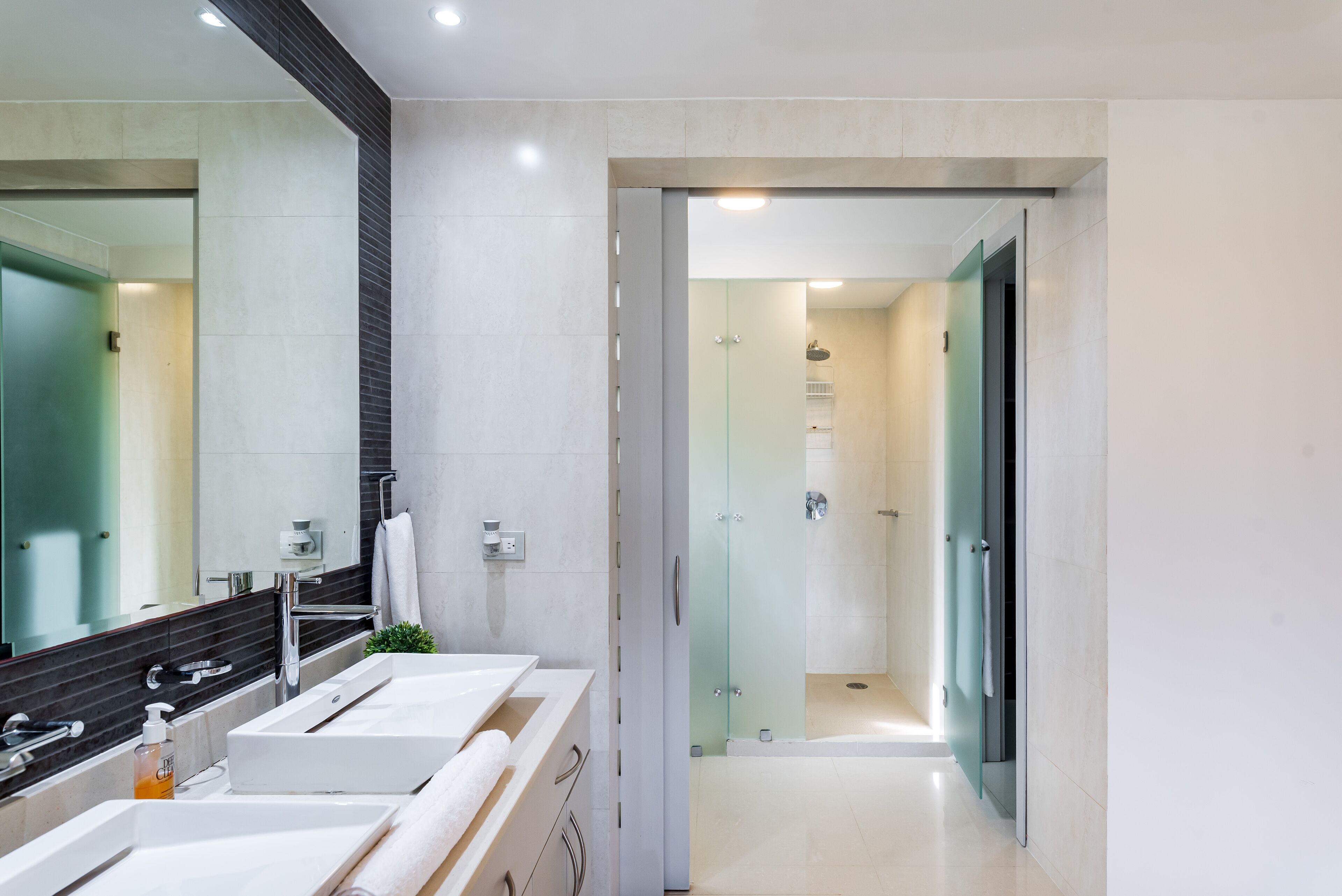 Une salle de bain moderne avec un grand miroir, deux lavabos et une douche séparée par une paroi de verre.