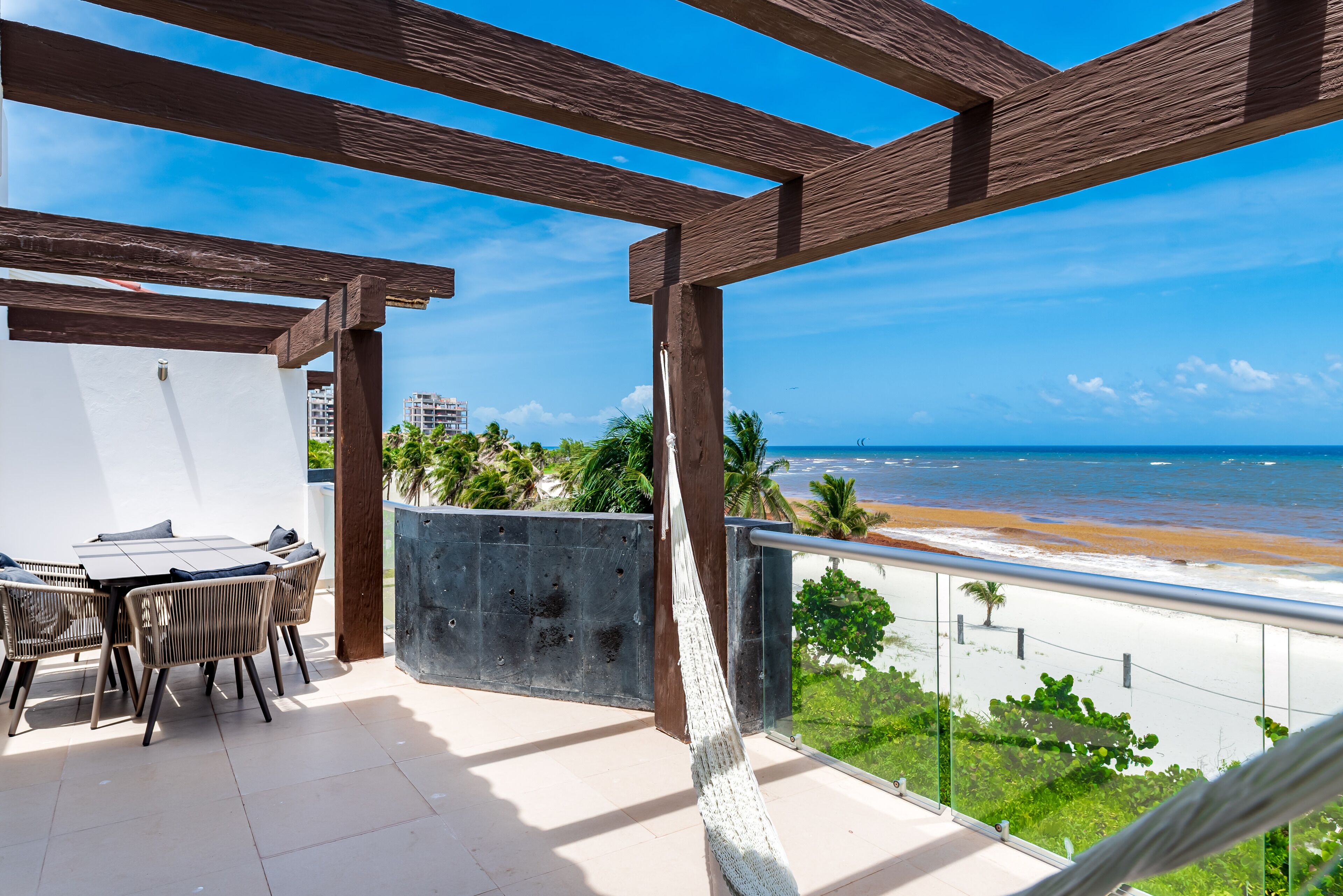 Un balcón con vista a la playa, una mesa con sillas y una hamaca.
