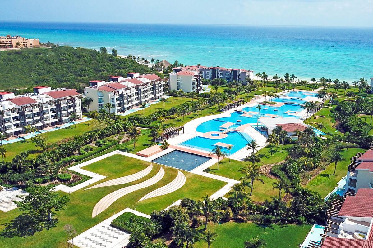 An aerial view of a resort with multiple swimming pools, lush greenery, and beachfront properties.