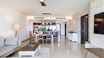 Een moderne woonkamer met een bank, salontafel en een eethoek. In de achtergrond is een keuken zichtbaar met een eettafel en stoelen.