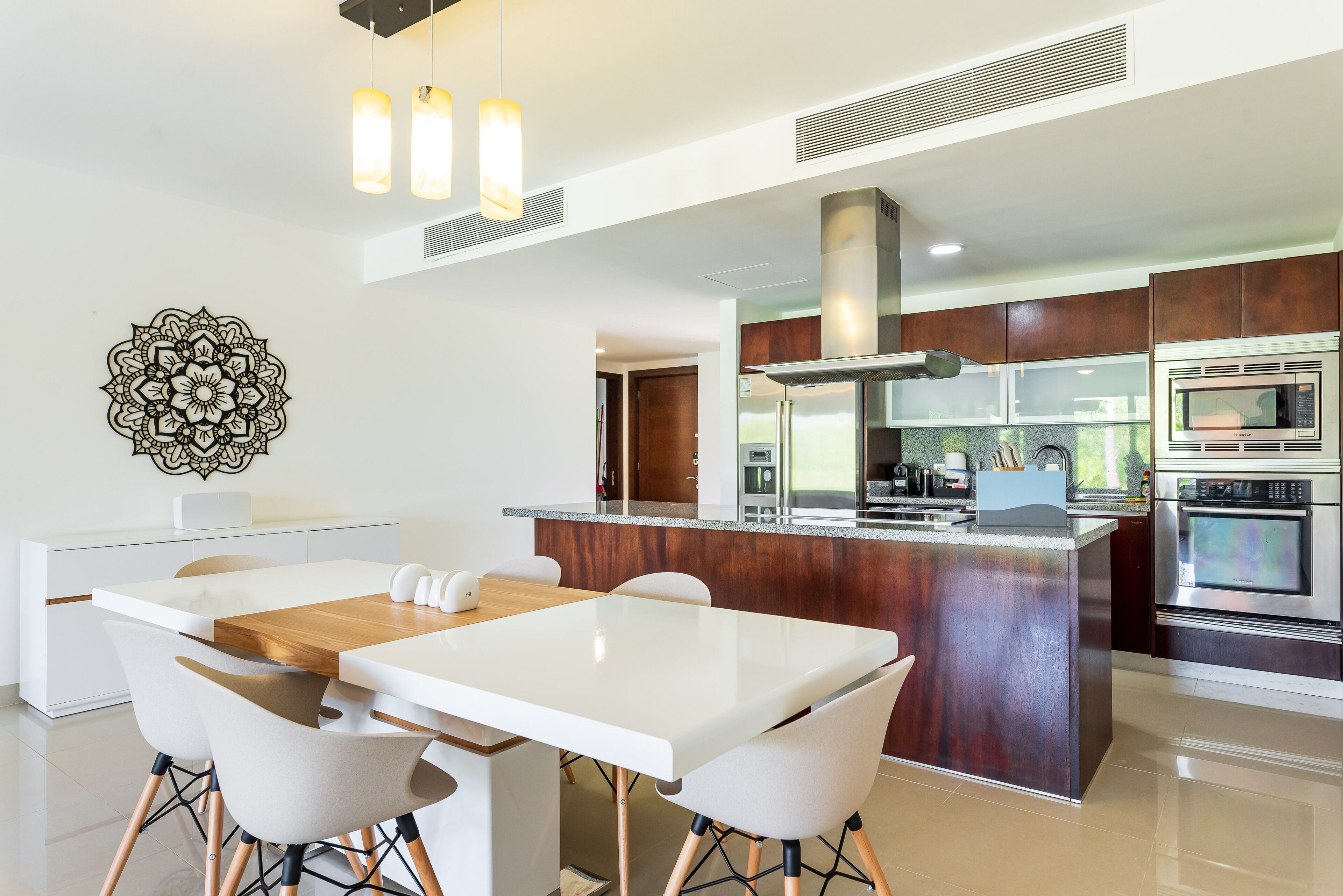 Uma cozinha moderna com uma ilha central, uma área de jantar com mesa e cadeiras brancas, e eletrodomésticos de aço inoxidável.