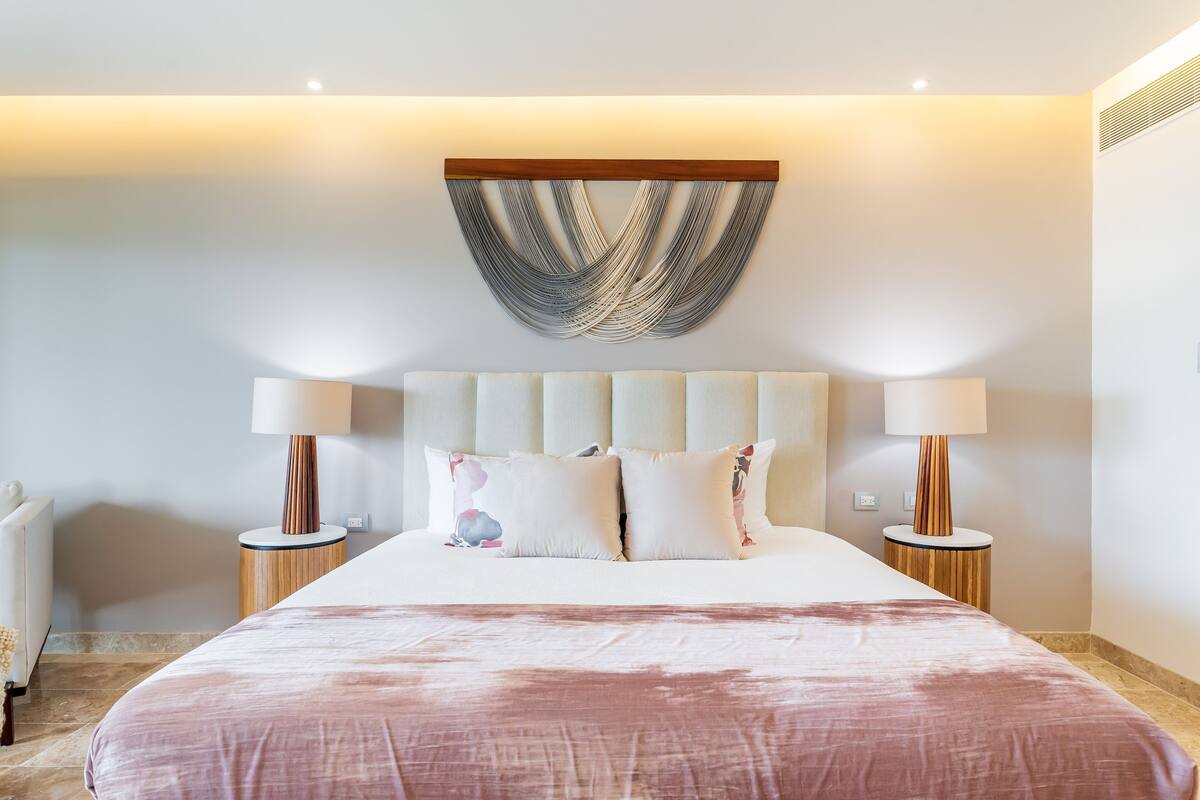 A modern hotel room with a large bed, two bedside lamps, a decorative wall hanging, and a radiator.