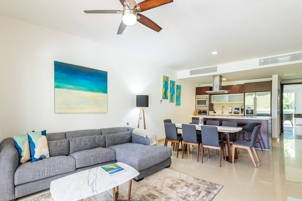 A modern living room with a grey sofa, a coffee table, and a ceiling fan. There is a dining area with a table and chairs, and a kitchen with a refrigerator and microwave.
