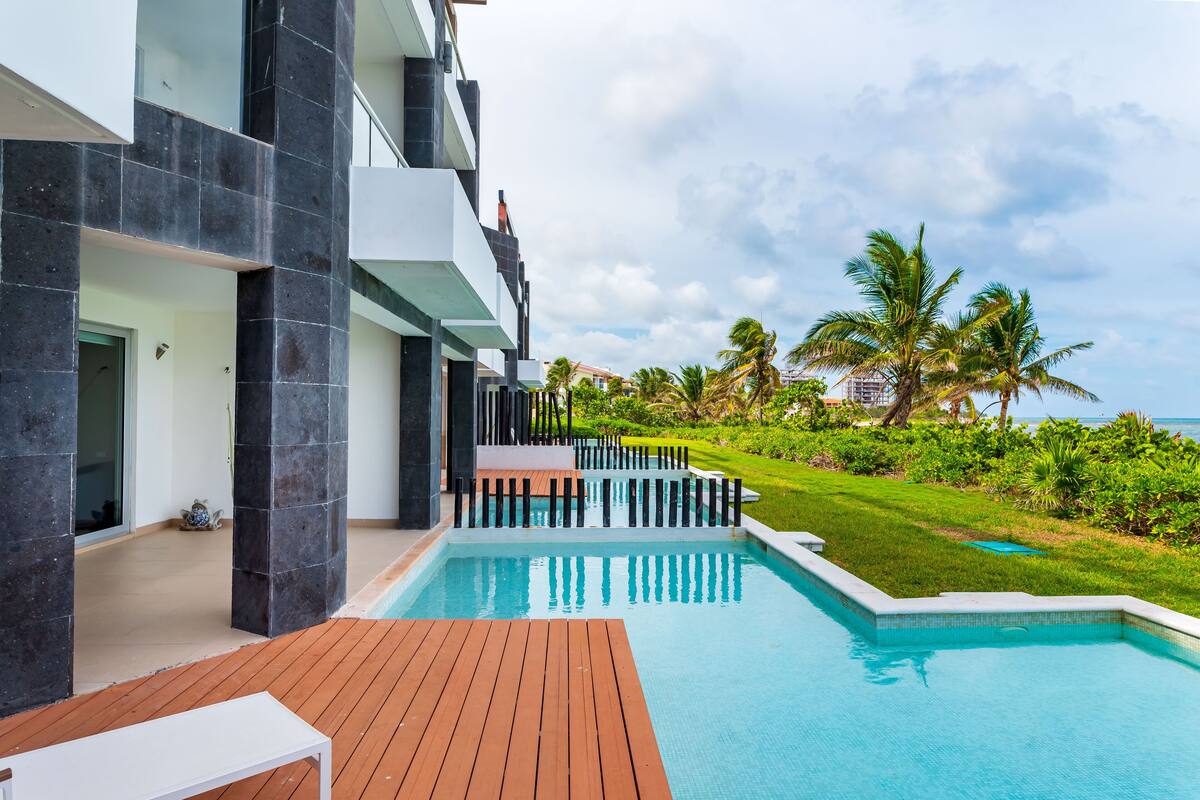 A modern hotel with a swimming pool, wooden deck, and a view of the ocean and palm trees.