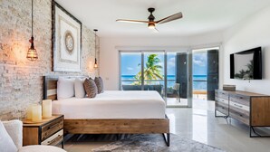 A modern bedroom with a large bed, a wooden nightstand, a desk, and a view of the ocean through a sliding glass door. - Mareazul Beach Condos Playa Del Carmen (Playa del Carmen)