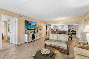 A spacious living room with a large sofa, a coffee table, and a television. There is a dining area in the background with a chandelier.