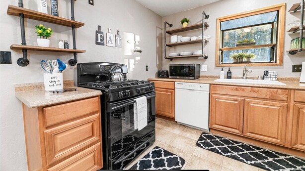A modern kitchen with wooden cabinets, a black stove, a white dishwasher, and a microwave. There are shelves with kitchen items and a window with a view.