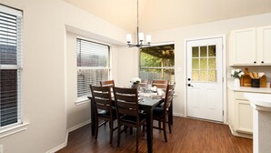 A dining area with a table set for four, surrounded by wooden chairs, a chandelier, and a view through a window with blinds.