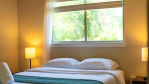 A hotel room with a bed, two bedside lamps, a window with a view of greenery, and a nightstand.