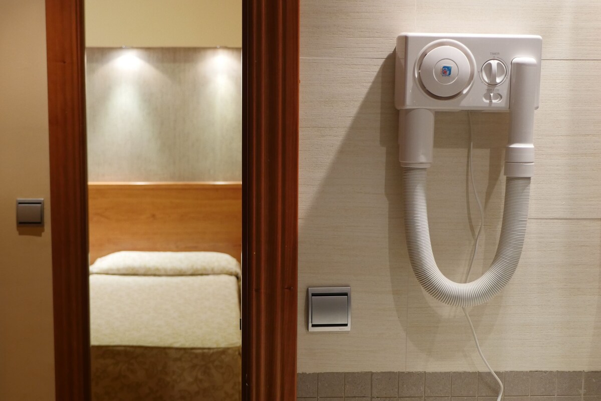 Una habitación de hotel con una puerta de madera, una cama con colchón blanco, un calentador fijado a la pared y un equipo de aire acondicionado instalado en la pared.